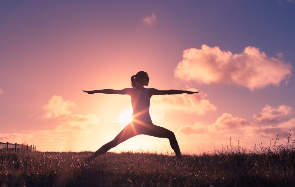 A woman does the "warrior" yoga pose in front of a sunset.