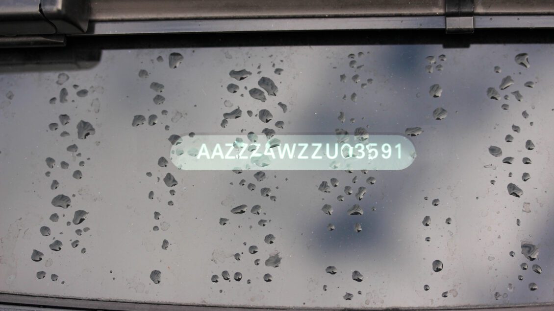 A VIN number is pictured in white writing affixed to the back windshield of a car splattered with rain.