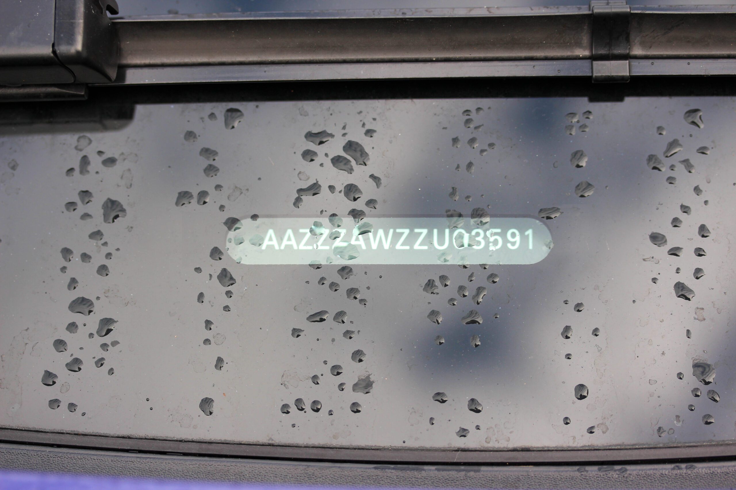 A VIN number is pictured in white writing affixed to the back windshield of a car splattered with rain.