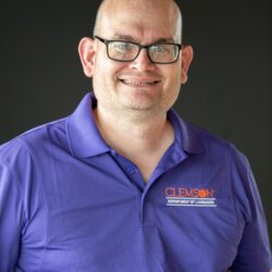 A man, Jason Hurdich, Senior Lecturer of American Sign Language, is balding and wearing glasses, a golf shirt. He has a shaved head and is smiling.