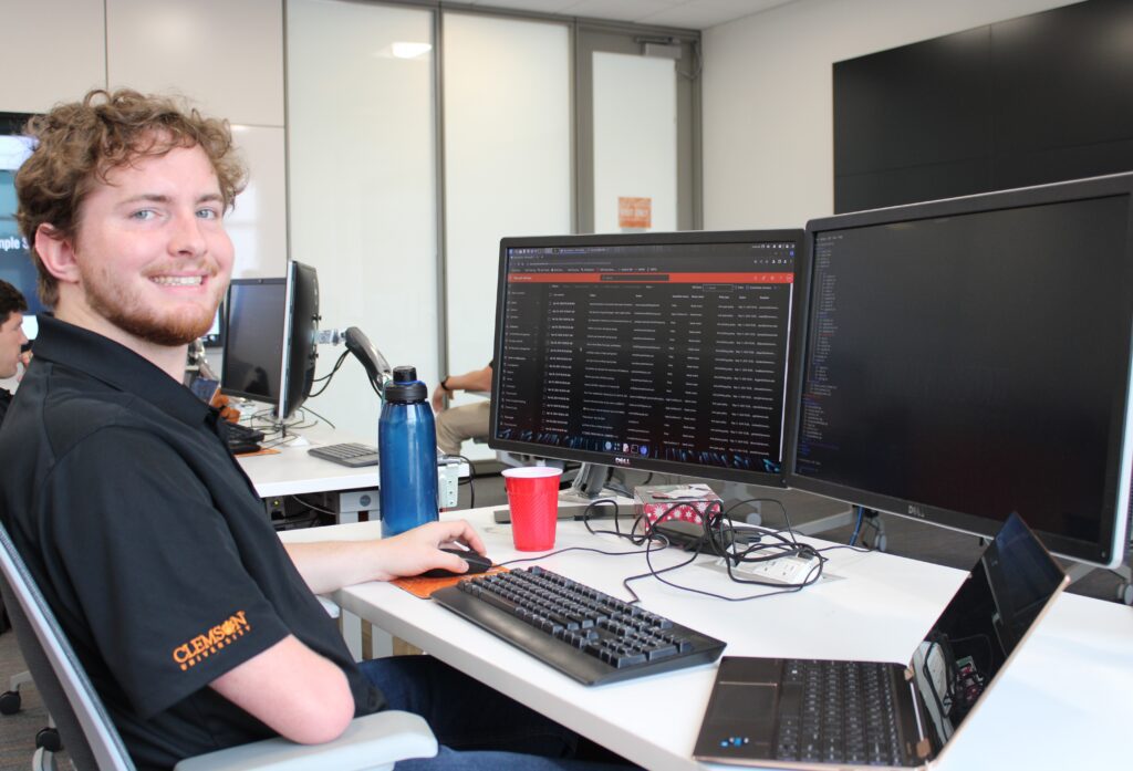 Duncan Hogg smiles while sitting in front of his desk with a laptop and two computer screens.