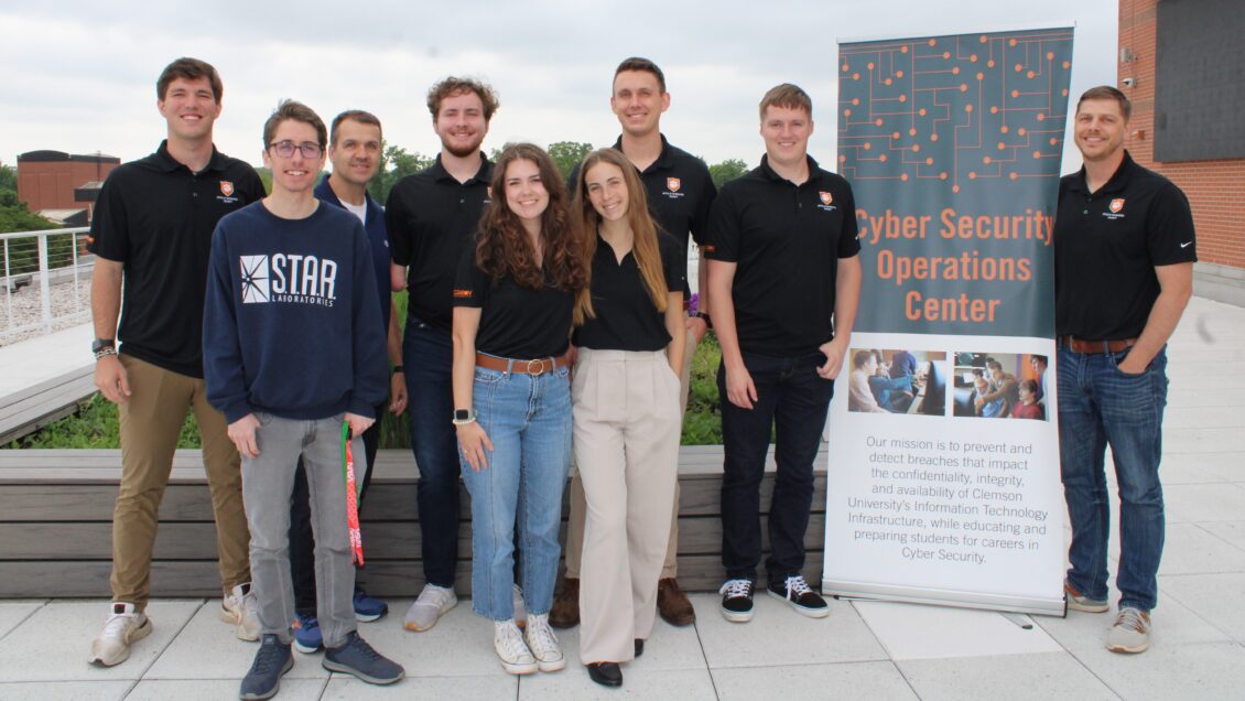 a group of students, CISO John Hoyt and CSOC Manager Calvin Watts stand on the roof of the Watt Center with a sign that says Cybersecurity Operations Center