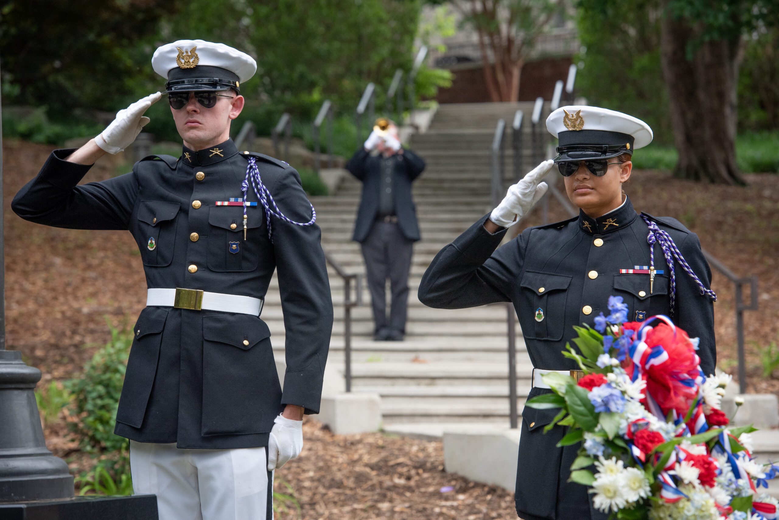 Two cadets in dress military uniforms salute while a trumpet player plays behind them