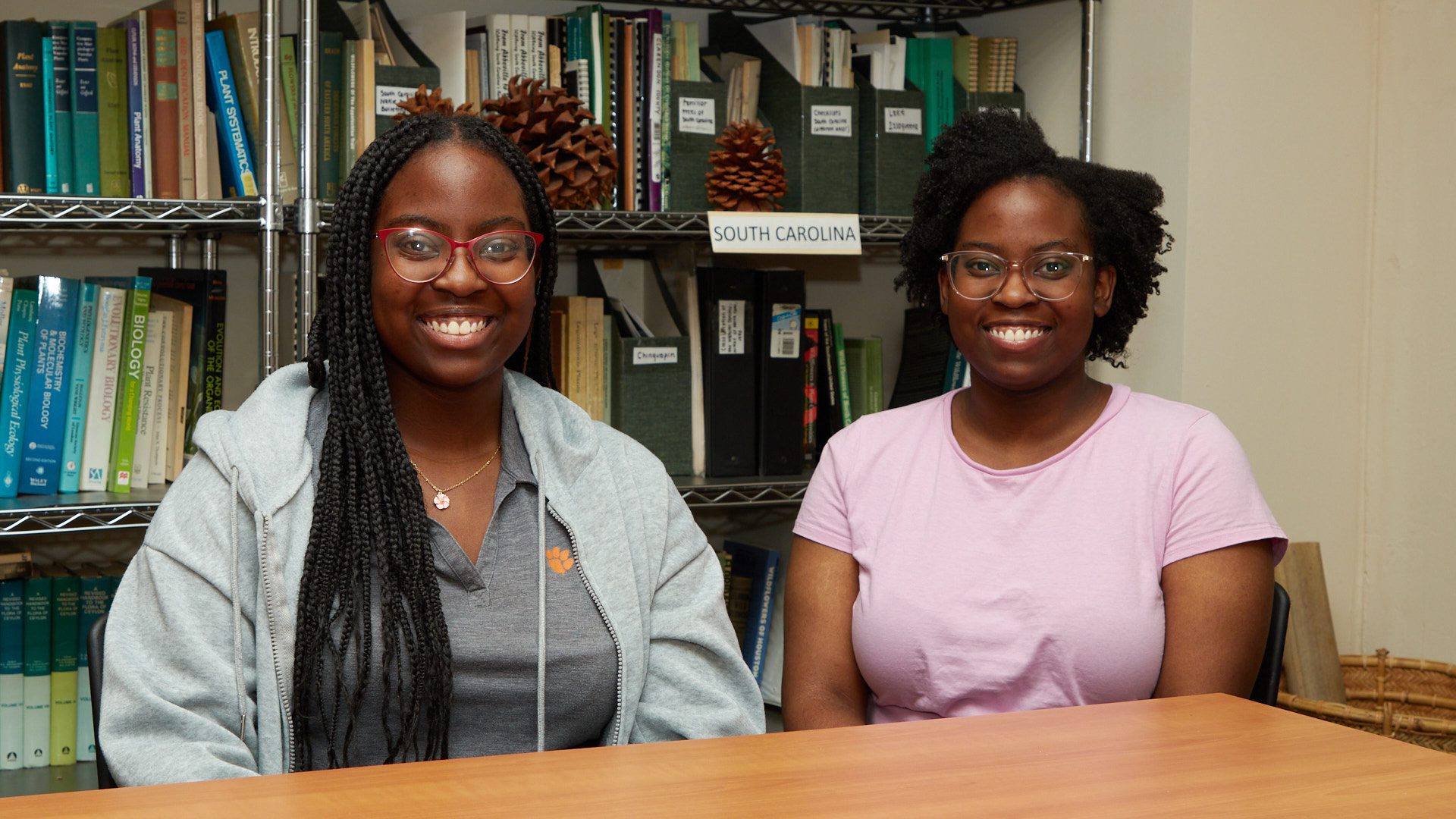 Two Black women are sitting at a table with a bookshelf full of books behind them.