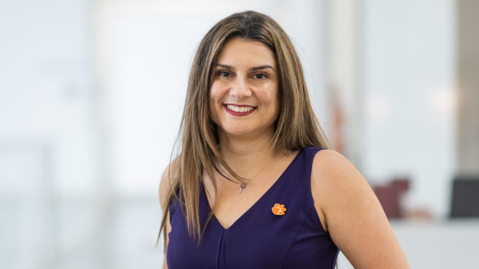 A woman with long hair wears a purple sleeveless shirt and an orange tiger paw pin and is smiling and facing forward.