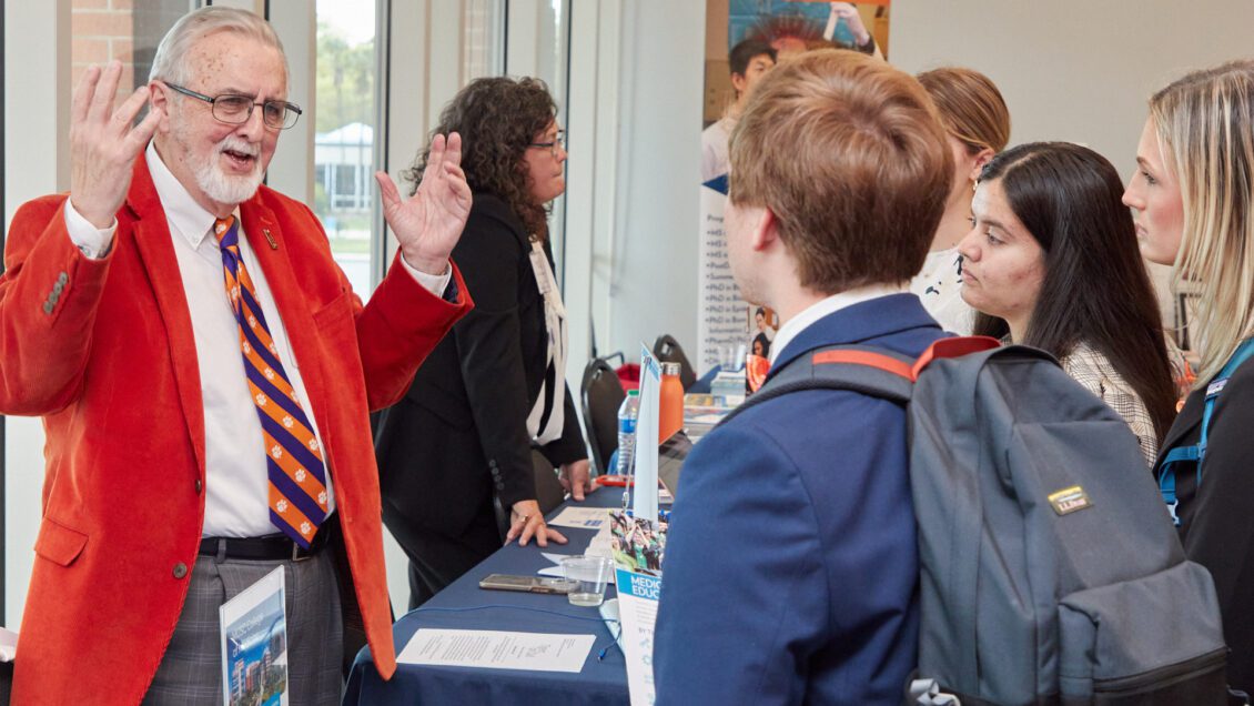 A man wearing an orange sports coat gestures to a student wearing a backpack that he's talking to at a career fair.