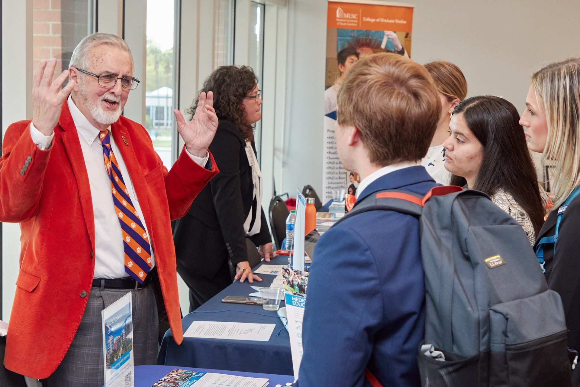 A man wearing an orange sports coat gestures to a student wearing a backpack that he's talking to at a career fair.