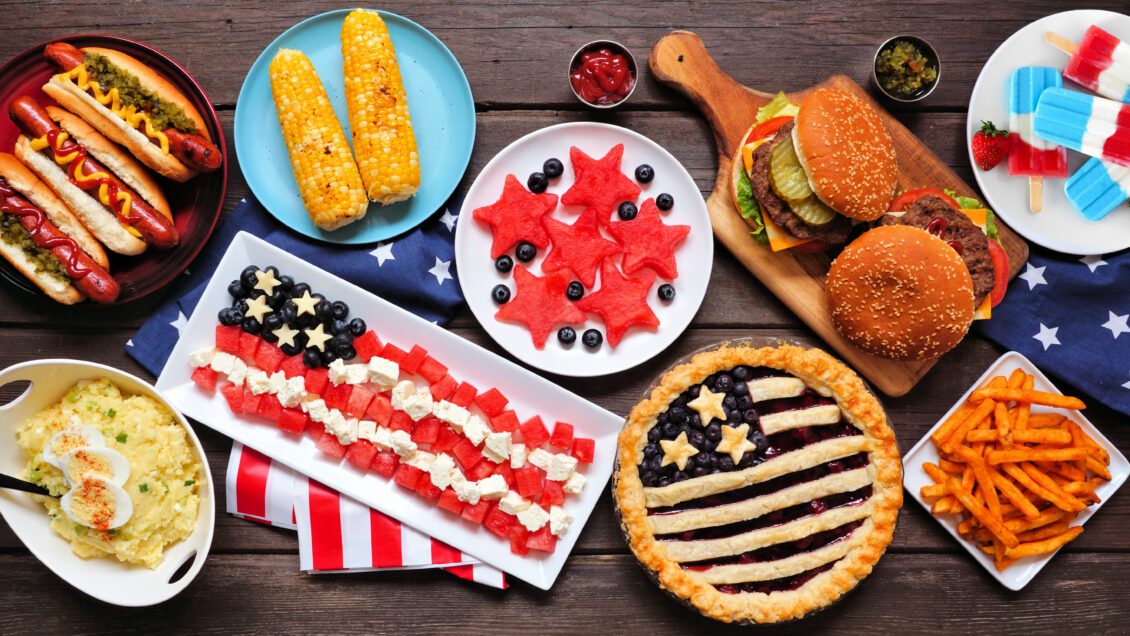 Assorted summer food such as pies, hot dogs and corn laid out on a table.
