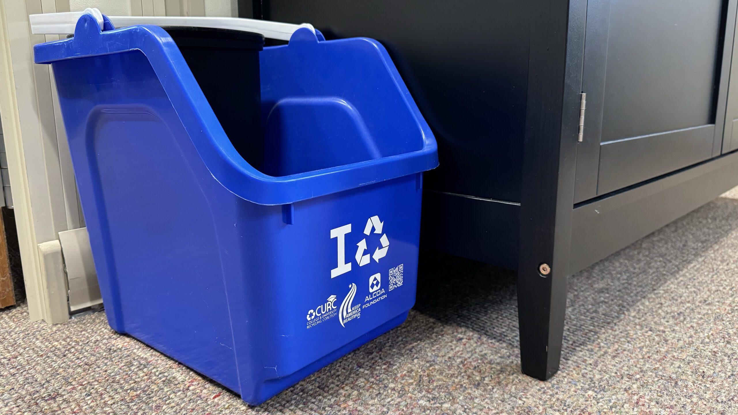 A small blue recycling bin on brown carpet next to a large black cabinet.