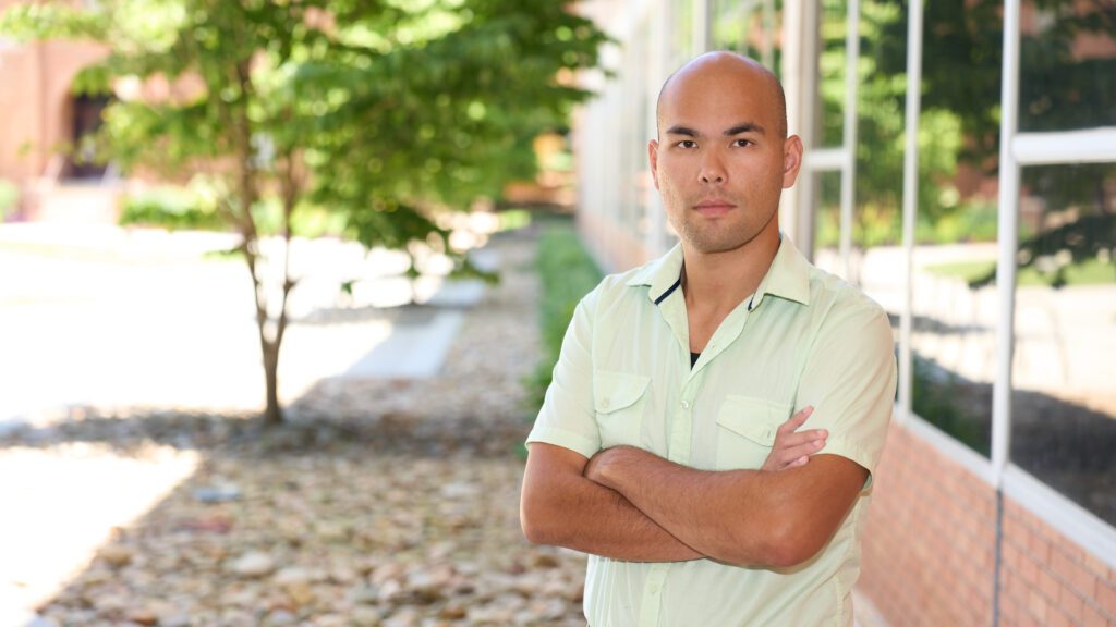 A man wearing a light colored shirt stands with his arms crossed in front of a building on the Clemson campus