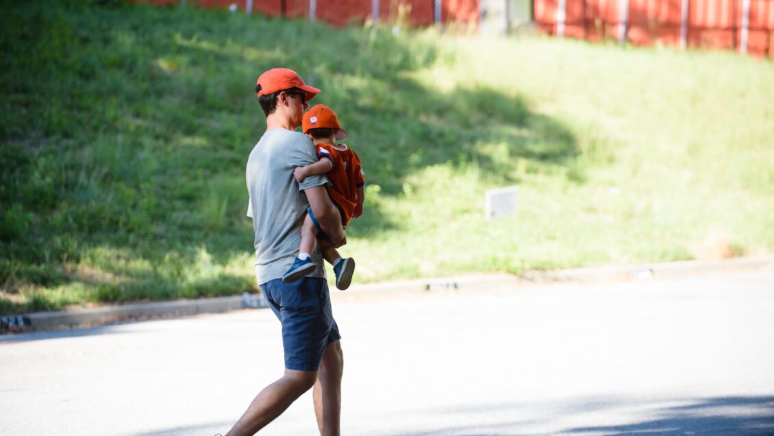 A man in a grey shirt and orange hat carries a toddler wearing a Clemson shirt and hat.