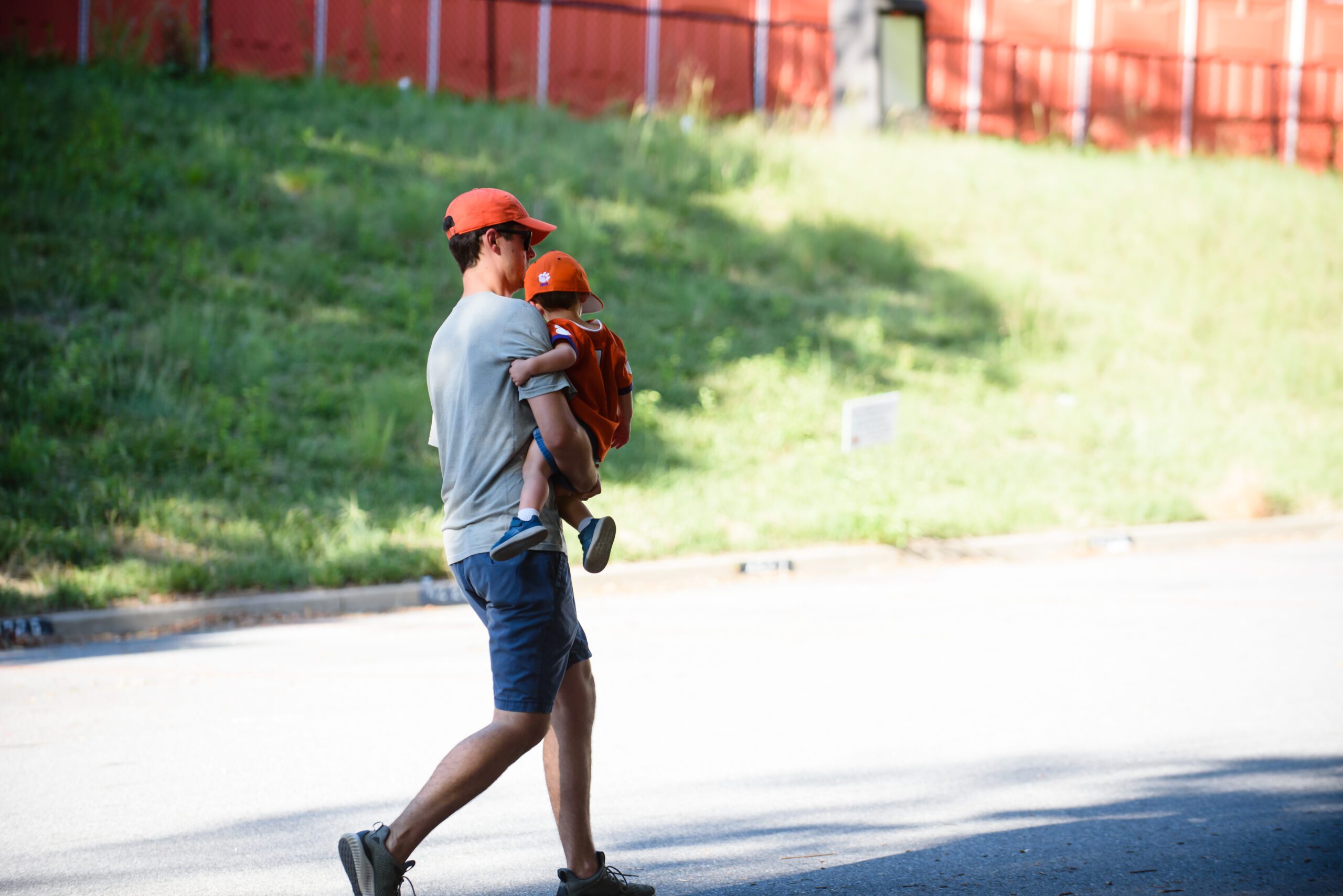 A man in a grey shirt and orange hat carries a toddler wearing a Clemson shirt and hat.