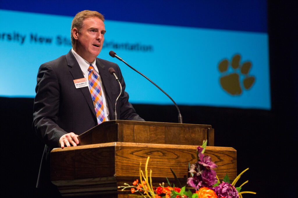 Caucasian man is a suit speaks while standing at a podium. Flowers are sitting in front of the podium, and a screen is behind the speaker.