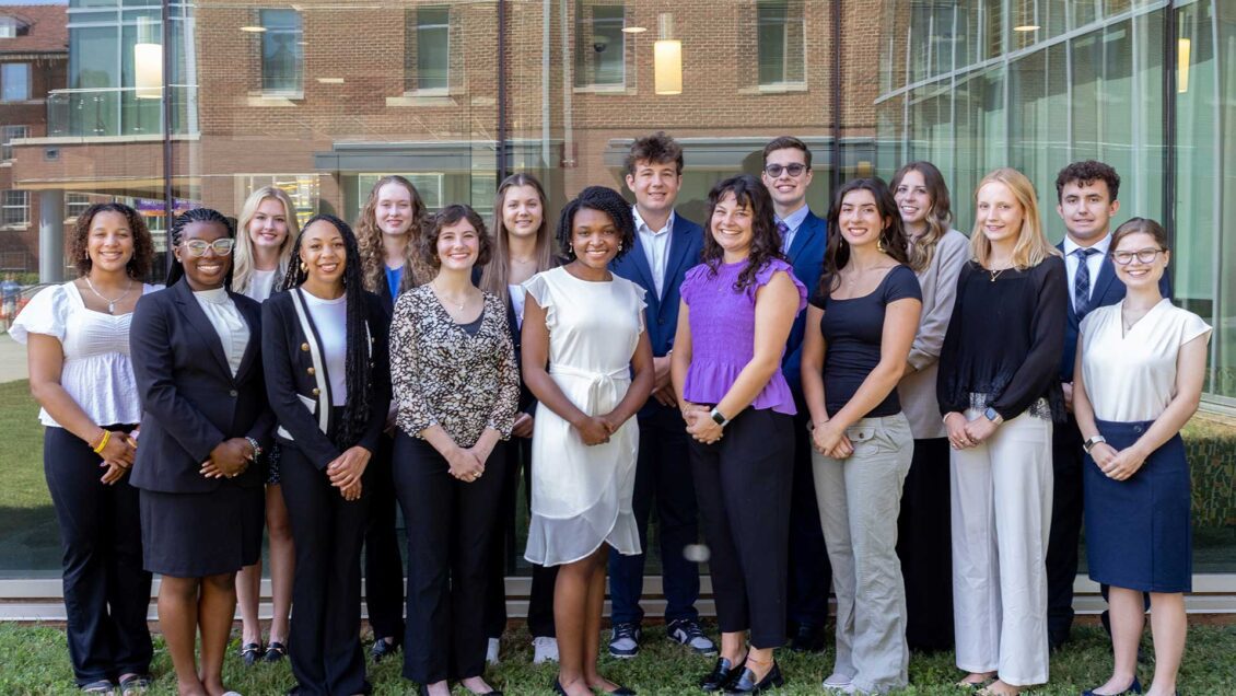 16 students standing outside in front of a window at the Honors Center on Clemson University campus.