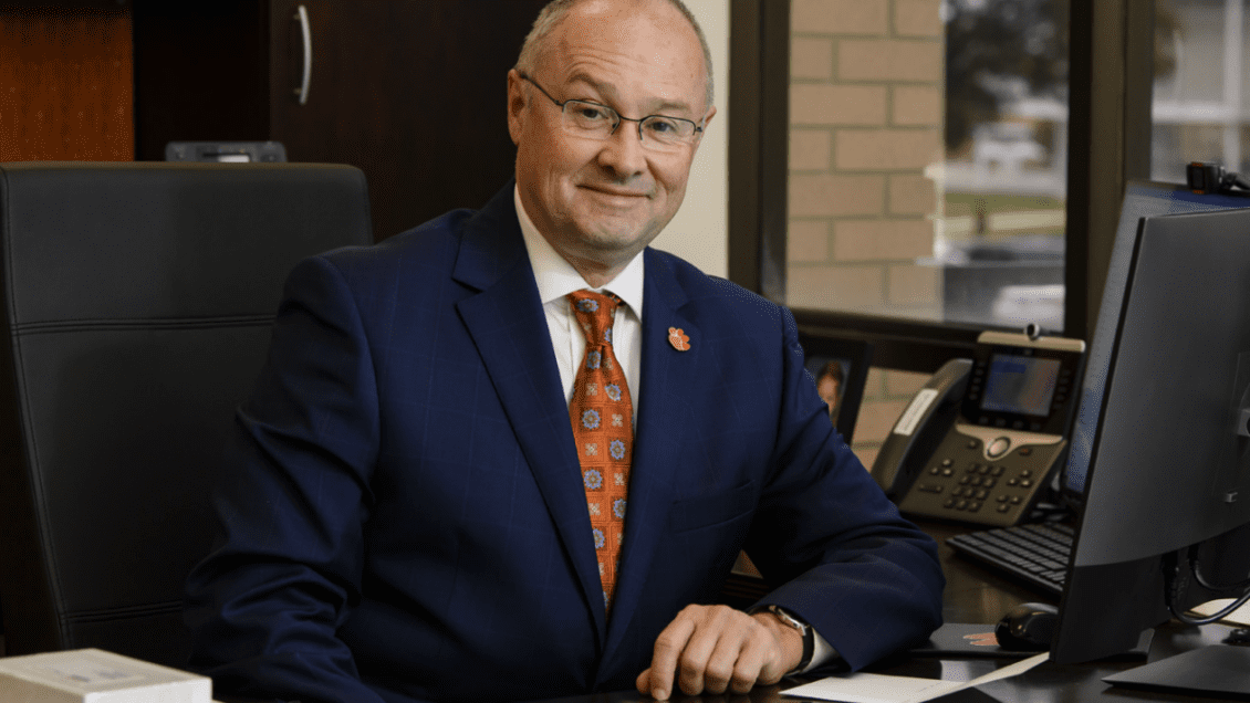 Nicholas Vazsonyi sits at a desk and smiles. He is wearing a suit and orange tie.