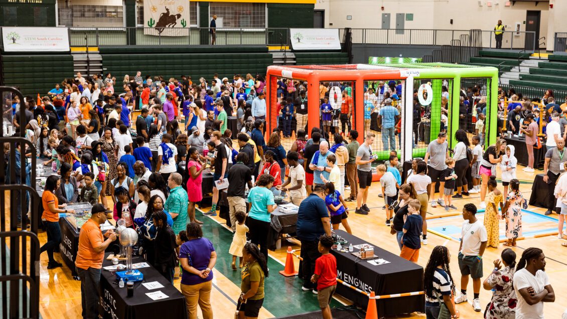 A crowded gym of K-12 students and adults, observing and participating in many STEM projects.