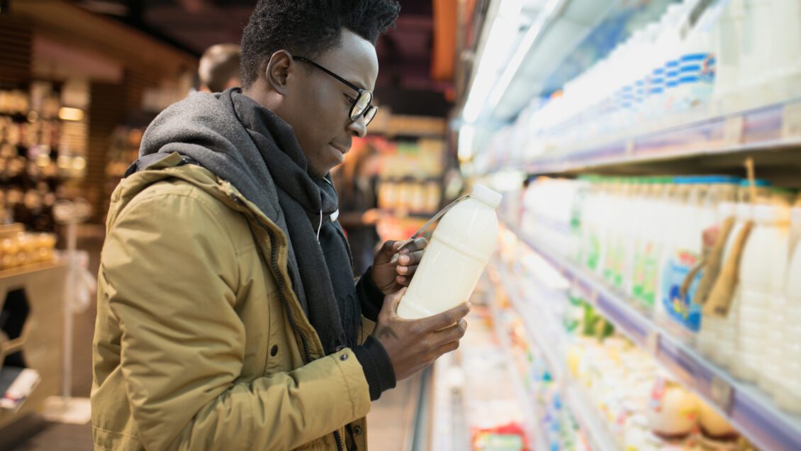 A young man wearing a coat and glasses stands in front of a grocery store display and is reading the back of a bottle.