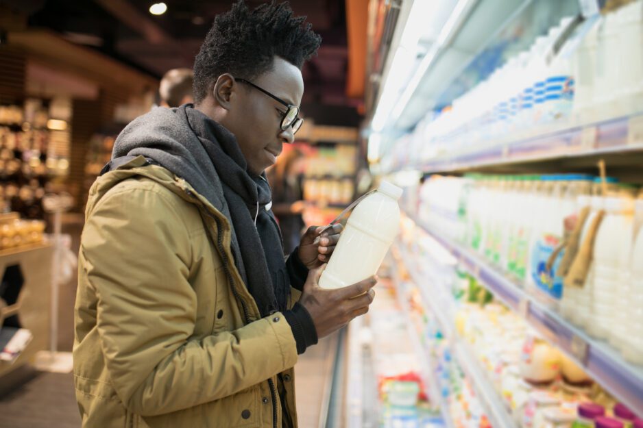 A young man wearing a coat and glasses stands in front of a grocery store display and is reading the back of a bottle.