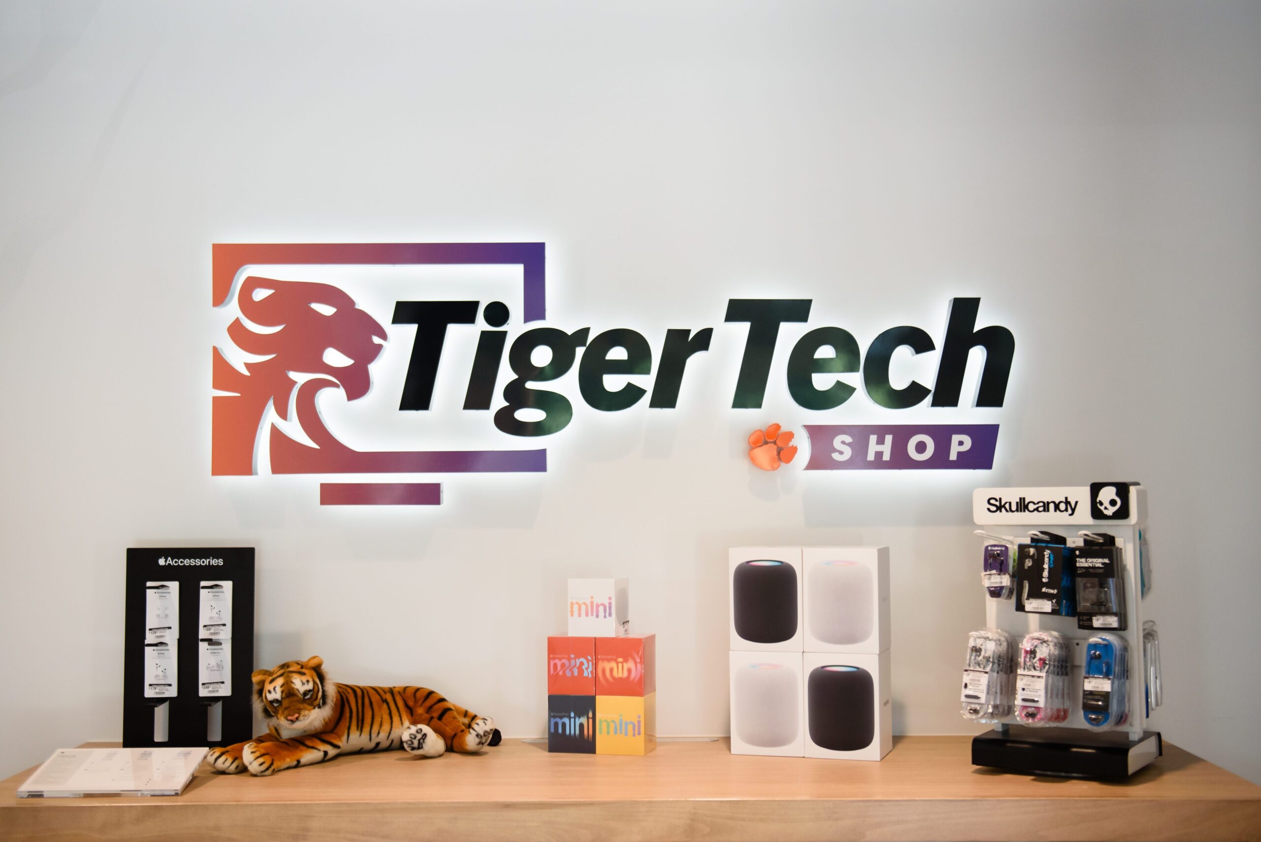 The Tiger Tech Shop logo is backlit hanging on a wall surrounded by Apple home pod products, Skull Candy accessories, and a small stuffed tiger.