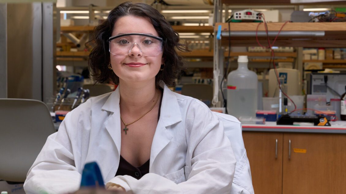 A woman wearing a white lab coat sits at the bench in a science lab