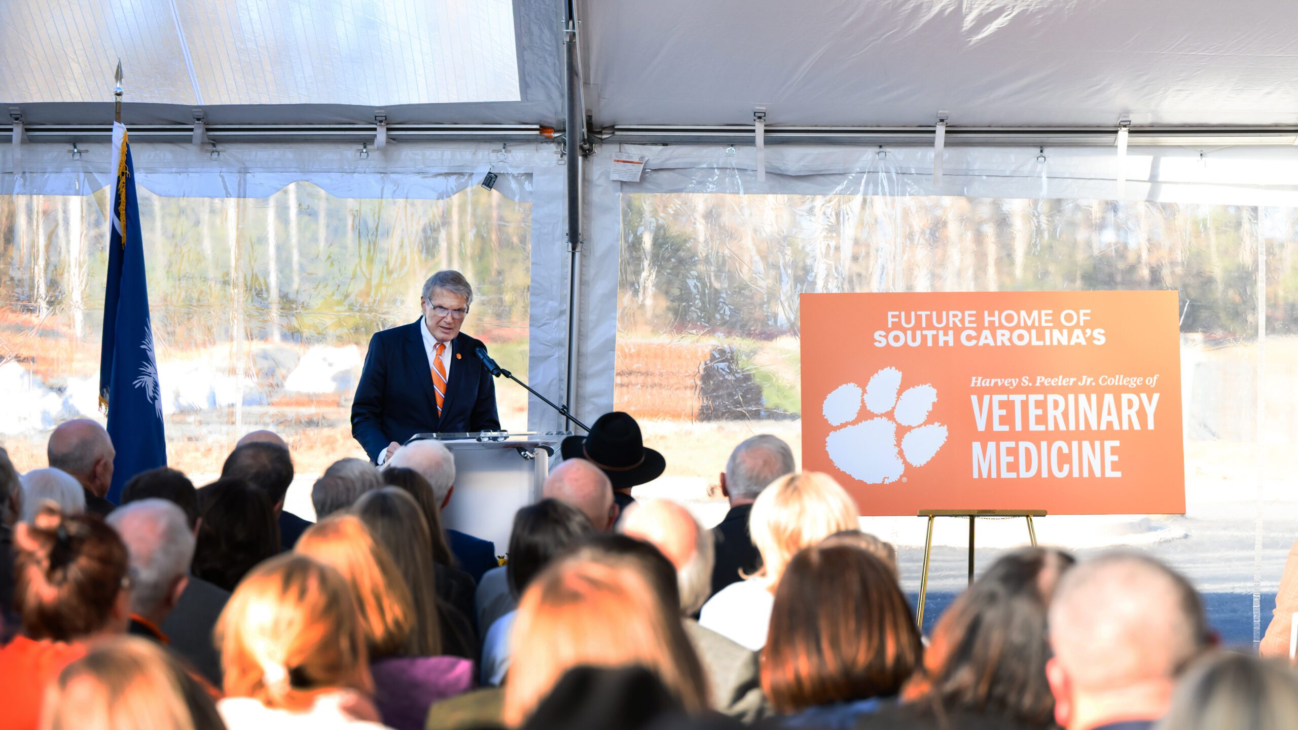 A man speaks at a podium while standing next to a Clemson sign while a seated audience watches.