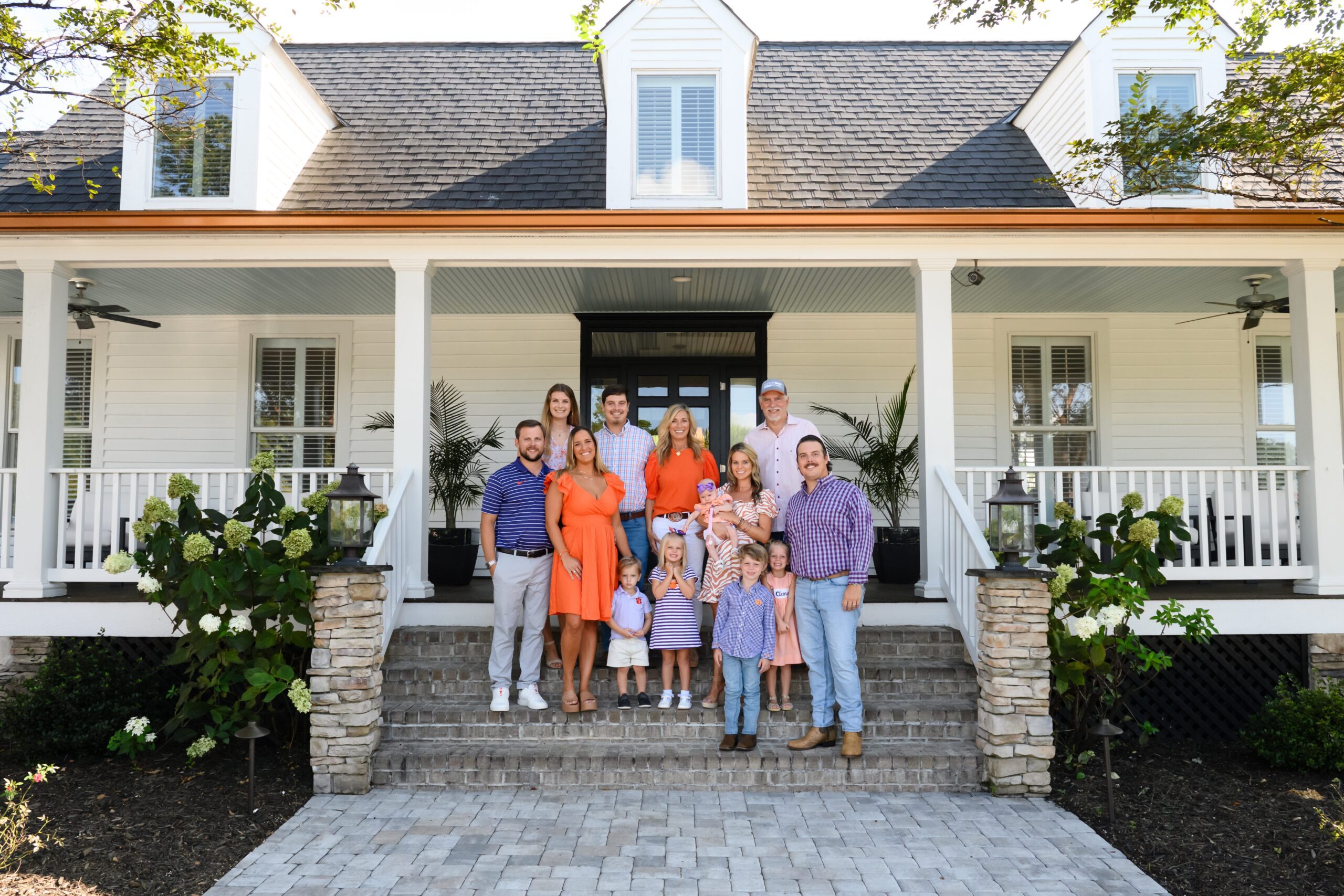 There generations of a family (Nieri) standing on steps leading to a wide porch of a large house. Two parents, three children and their spouses and five grandchildren (from a baby to elementary school ages).