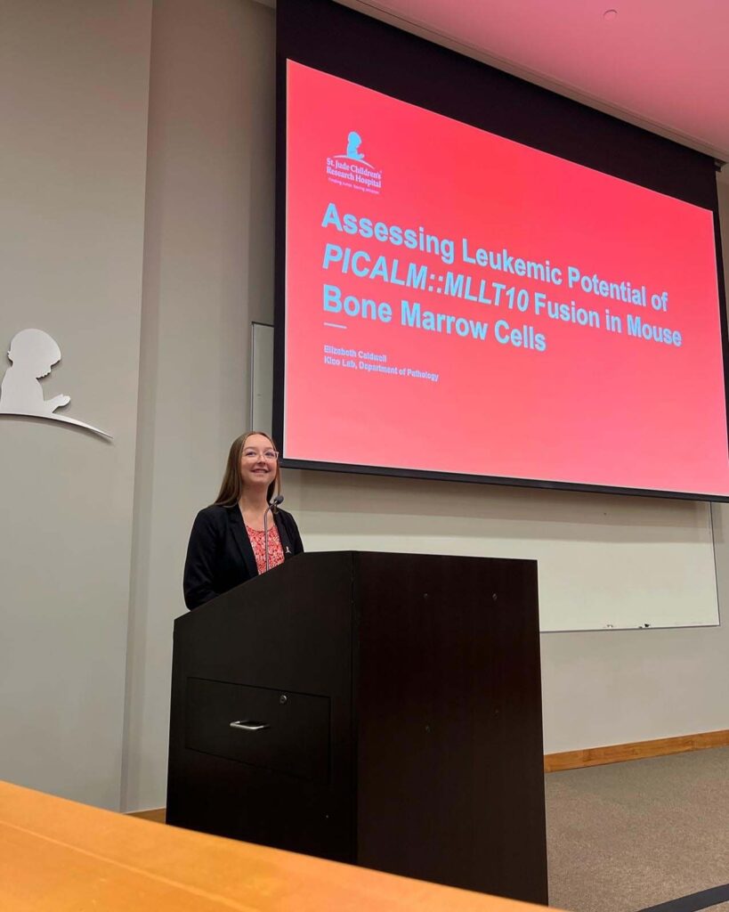 A young woman stands behind a large black podium on a stage with a big screen above her left shoulder that reads, "Accessing Leukemic Potential of PICALM:: MLLT10 Fusion in Mouse Bone Marrow Cells