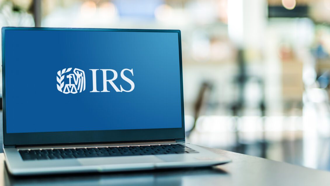 A black laptop computer sits on a desk and is open to a screen with the blue IRS logo on it.