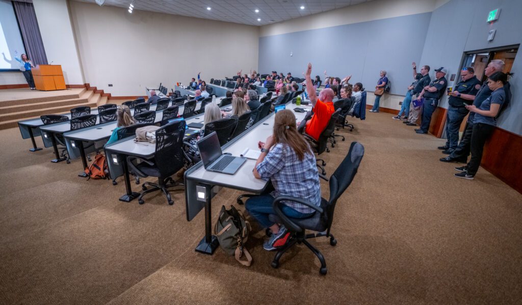 A room full of people sit in rows of desks with some raising their hands