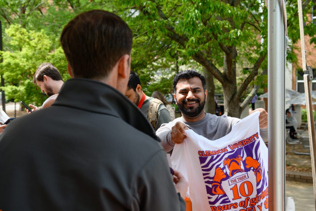 Male student with a smile, beard and dark complexion holds up a 10th annual give day t-shirt that features The Tiger. He is chatted at an outdoor booth.