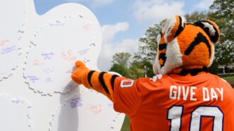 The Tiger is pointing to signatures on a six foot tall tiger paw cut out that has donor signatures. The Tiger is wearing a football jersey with Give Day and the number ten on the back.