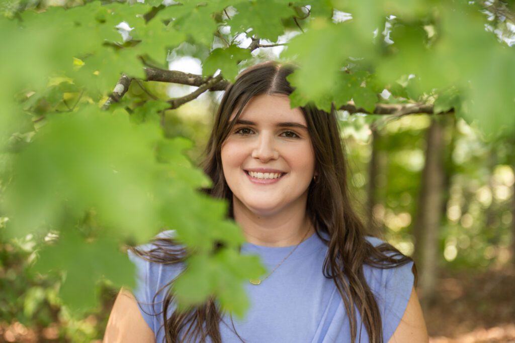 A young woman with long brown hair wearing a lavender shirt smiles at the camera through leaves in the forefround.