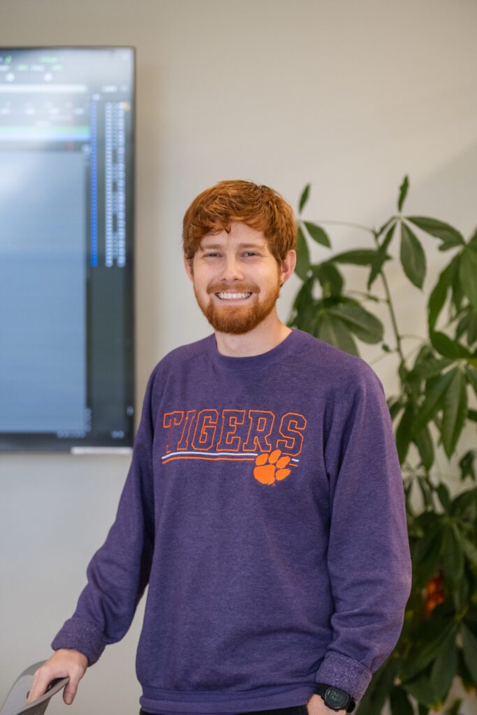A young man with red hair and facial hair stands and smiles at the camera wearing an orange sweatshirt that says "Tigers."