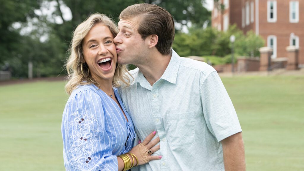 A young man kisses the cheek of a young woman while they stand in a grassy area with brick buildings in the background. They are siblings.
