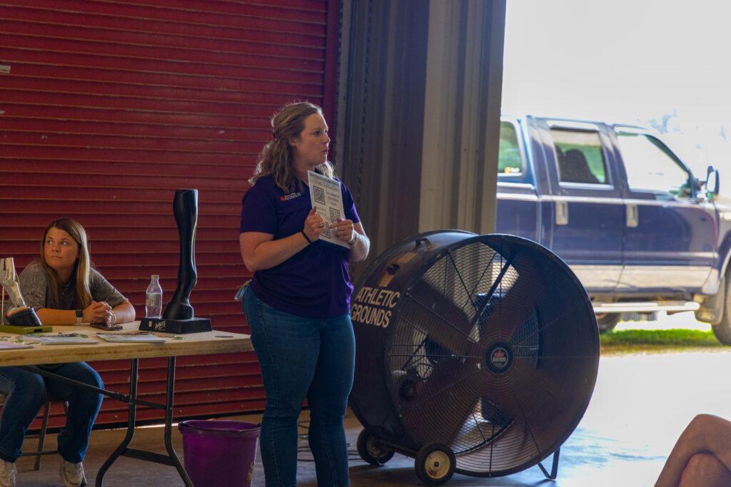 A woman stands in the doorway of a pole barn and speaks to an audience while pointing at a card she's holding.