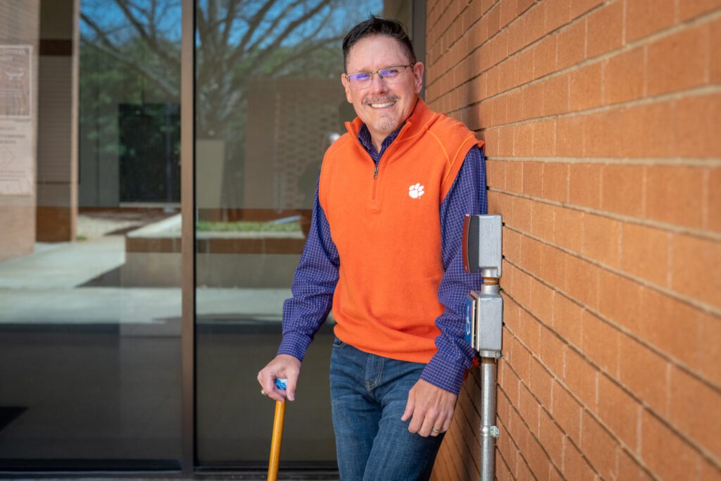 A man with a cane leans on a brick wall, smiling and wearing an orange Clemson vest, in front of a button that opens the automatic doors behind him.