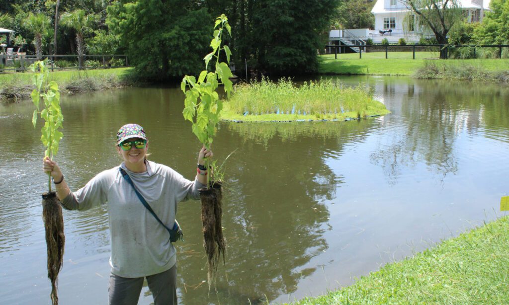 A woman wearing a hat and sunglasses holds up two plants with long, wet root systems with water in the background.