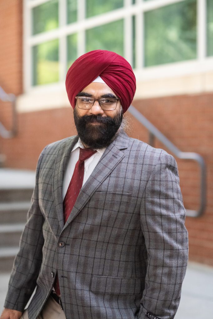 A bearded man wearing a plaid suit, red turban and glasses smiles at the camera at the bottom of a staircase in front of the exterior of a brick building.