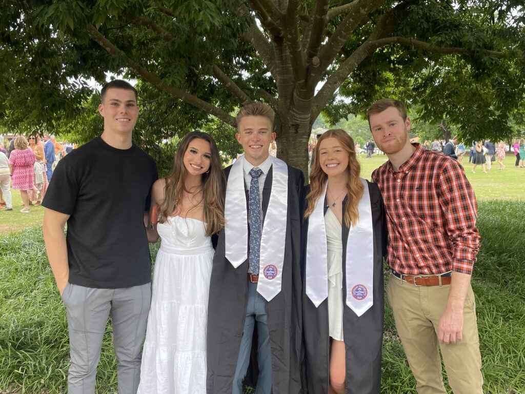 A group of five people stand under a tree with their arms around each other, two in the middle wearing graduation robes.