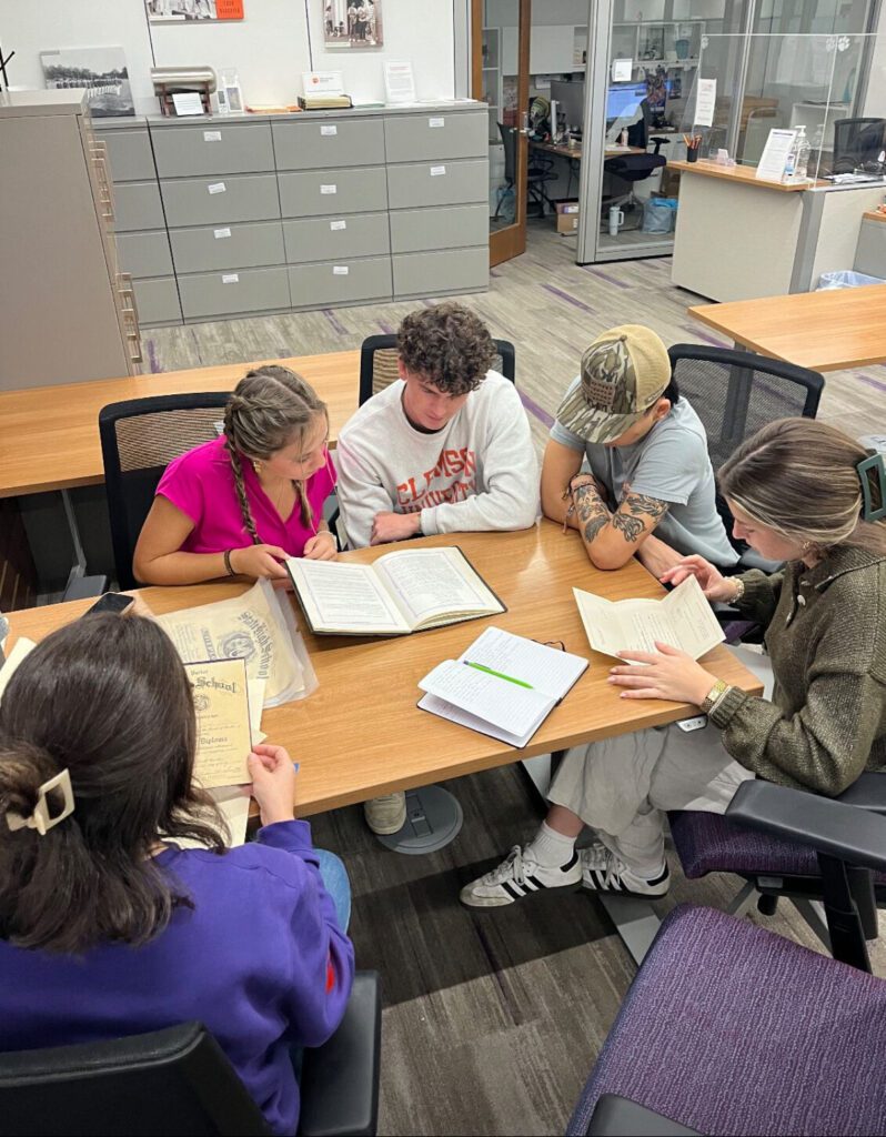A group of four students sit around the end of a table looking at books and documents.