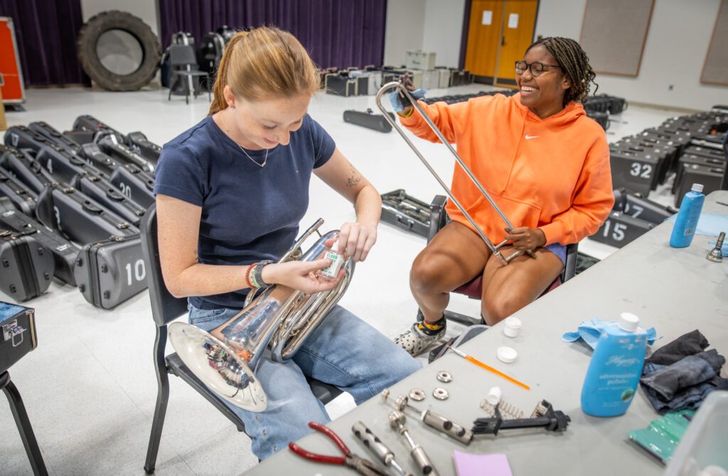 Two women sit at a desk and clean musical instruments - one has a baritone and one has a trombone slide.