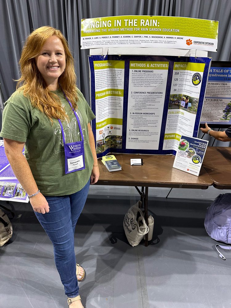 A woman wearing an event badge stands next to a table with a cardboard display on it titled "Racing in the Rain."
