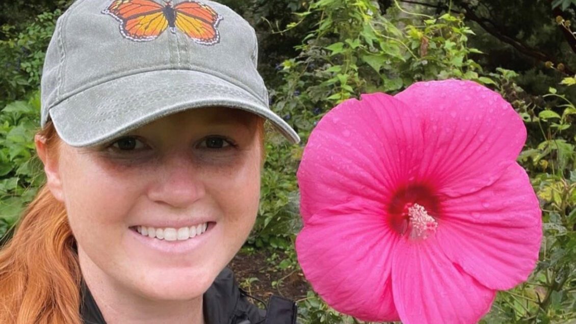 A woman with long red hair wearing a baseball cap with a butterfly on it stands next to a large pink flower that's as big as her head.