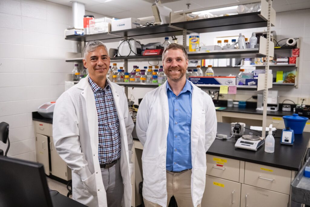 Two men in white lab coats are standing in a scientific lab smiling. 