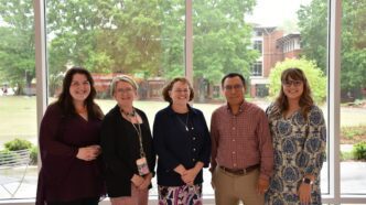 A group of five people representing the Clemson AI team are dressed in business casual attire and standing indoors in front of large windows with views of the campus behind them.