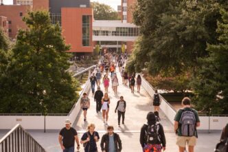 A large group of college students, many carrying backpacks, walks across the Library Bridge on the Clemson University campus. In the background, an academic building with glass and brick exterior walls is visible.