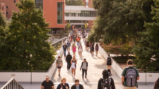 A large group of college students, many carrying backpacks, walks across the Library Bridge on the Clemson University campus. In the background, an academic building with glass and brick exterior walls is visible.