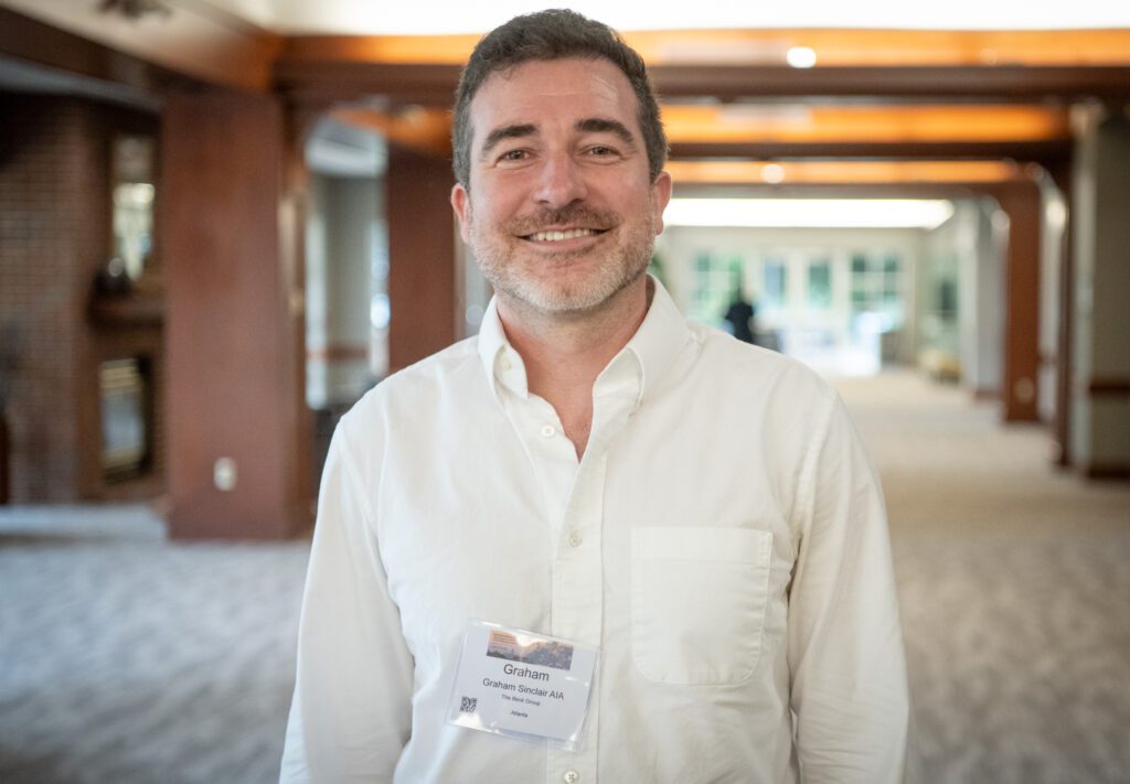 The man in a white collared shirt with straps around his neck stands in a wide hallway smiling at the camera.