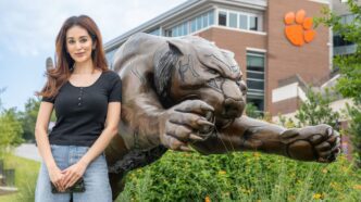 A woman with long dark hair leans on a large bronze statue of a tiger pouncing, its clawed paws reaching in front of it, with a Clemson Tiger paw on Memorial Stadium visible in the background.