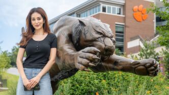 A woman with long dark hair leans on a large bronze statue of a tiger pouncing, its clawed paws reaching in front of it, with a Clemson Tiger paw on Memorial Stadium visible in the background.